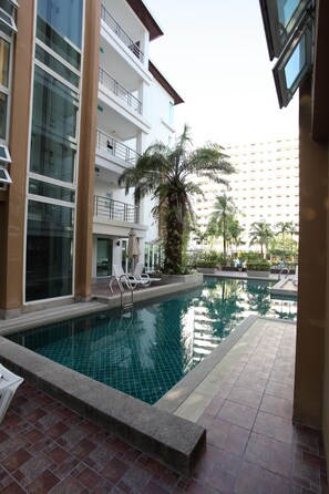 A swimming pool with lounge chairs, a palm tree, and a multi-story building in the background.
