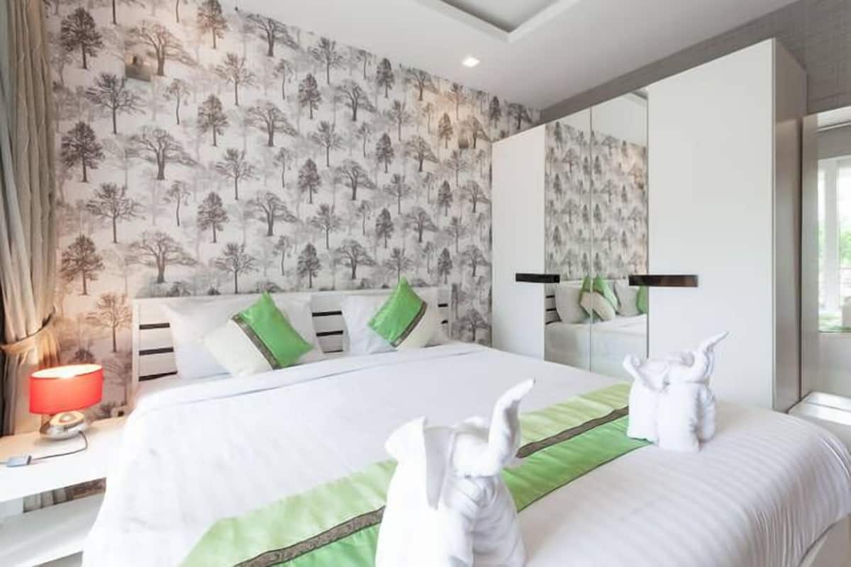 A hotel room with a large bed, green and white bedding, a red lamp, and a forest-themed wallpaper.