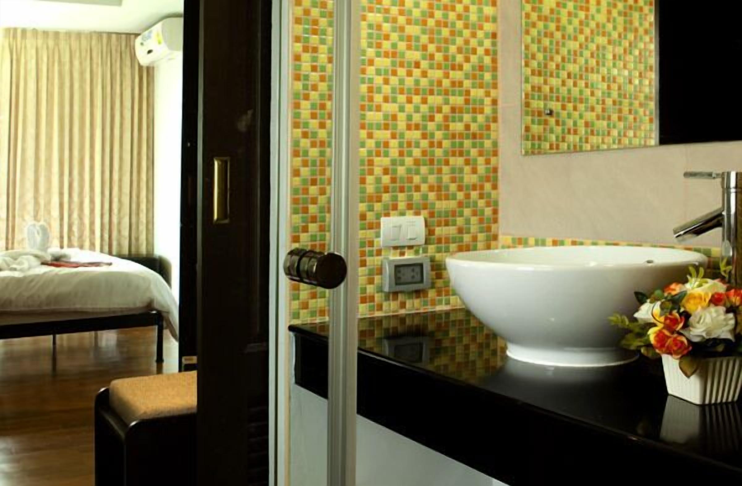 Habitación de hotel con un lavamanos grande de color blanco, una cama con edredón blanco y un baño con paredes de mosaicos.