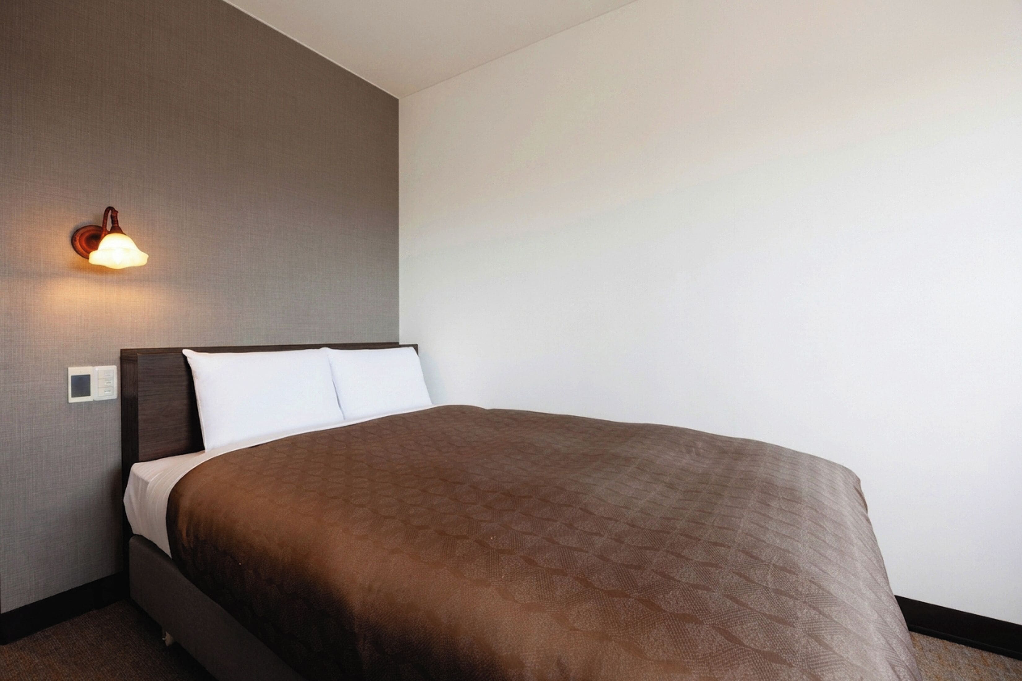 A hotel room with a bed, a wall-mounted light, and a thermostat.