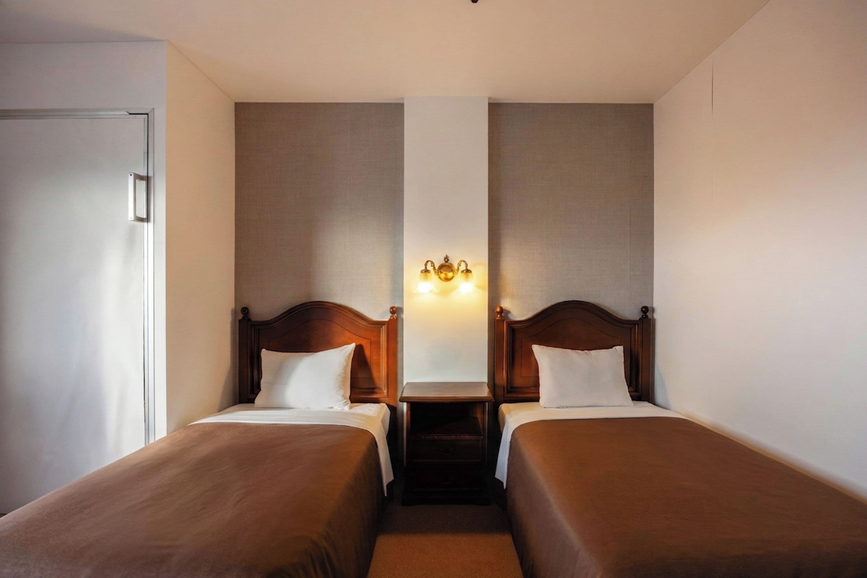 A hotel room with two single beds, a nightstand, a wall-mounted light fixture, and a door.