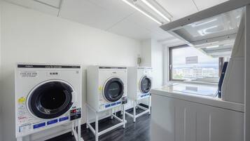 A laundry facility with three front-loading machines, a window with a view of buildings, and a white ceiling with fluorescent lights.