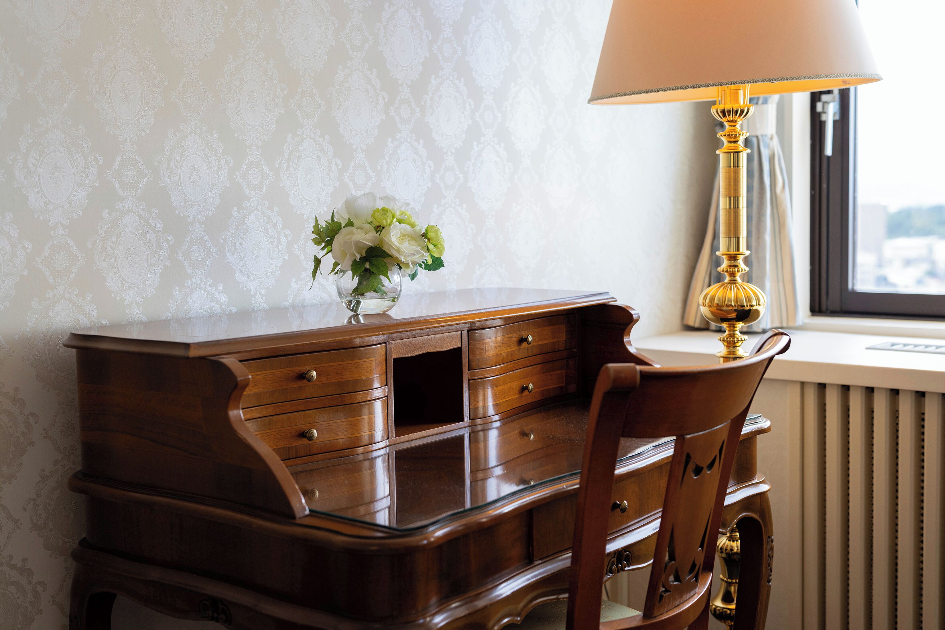 A wooden desk with drawers, a vase with flowers, a lamp, and a chair.
