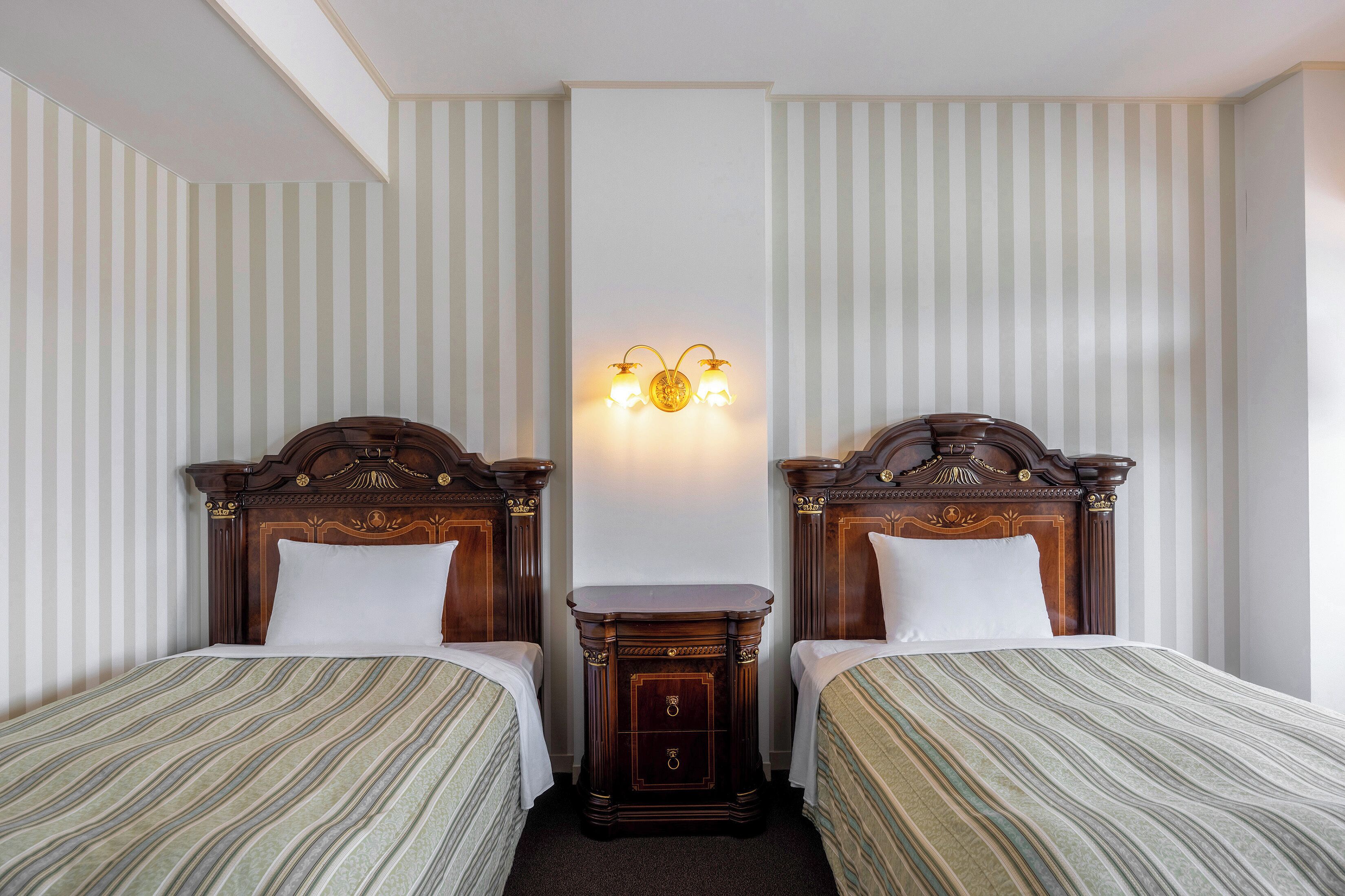 Habitación de hotel con dos camas individuales, una mesita de noche, una lámpara fijada en la pared y papel tapiz a rayas.