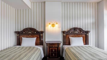 Habitación de hotel con dos camas individuales, una mesita de noche, una lámpara fijada en la pared y papel tapiz a rayas.