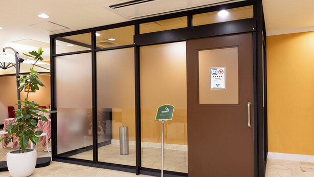 Entrada de hotel con puerta corredera de vidrio, una planta en maceta y señal de zona para fumadores.