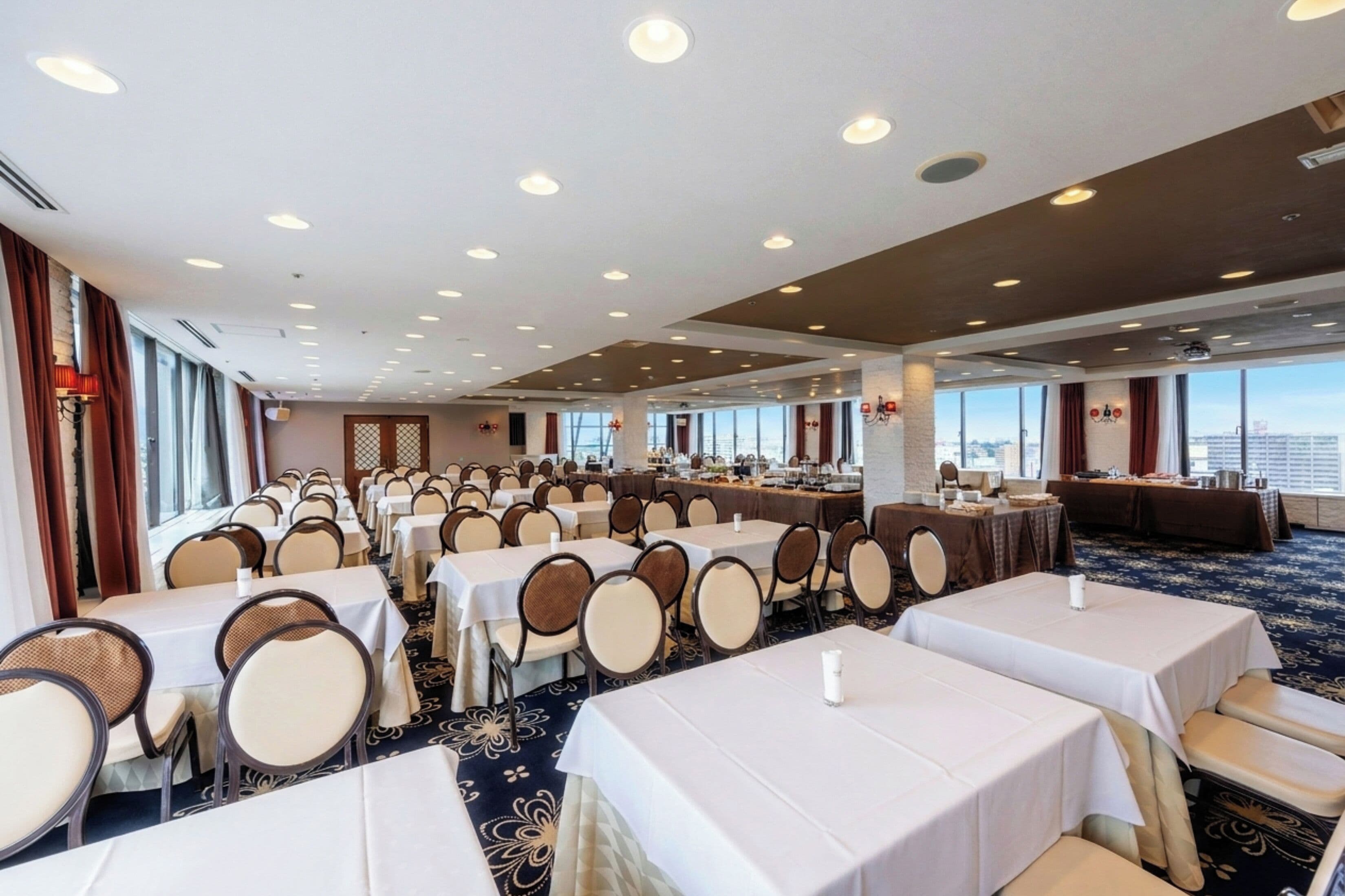 Un salón de conferencias de hotel con varias mesas y sillas, ventanas amplias y un techo alto con iluminación empotrada.