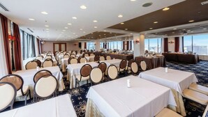 Un salón de conferencias de hotel con varias mesas y sillas, ventanas amplias y un techo alto con iluminación empotrada.