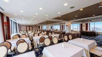 Un salón de conferencias de hotel con varias mesas y sillas, ventanas amplias y un techo alto con iluminación empotrada.