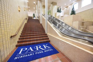 Entrada de un hotel con alfombra azul que indica PARK HIROSAKI PARK HOTEL, una escalera y una escalera mecánica adyacente.