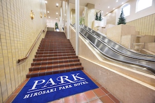 A hotel entrance with a blue carpet indicating the PARK HIROSAKI PARK HOTEL, a staircase, and an adjacent escalator.
