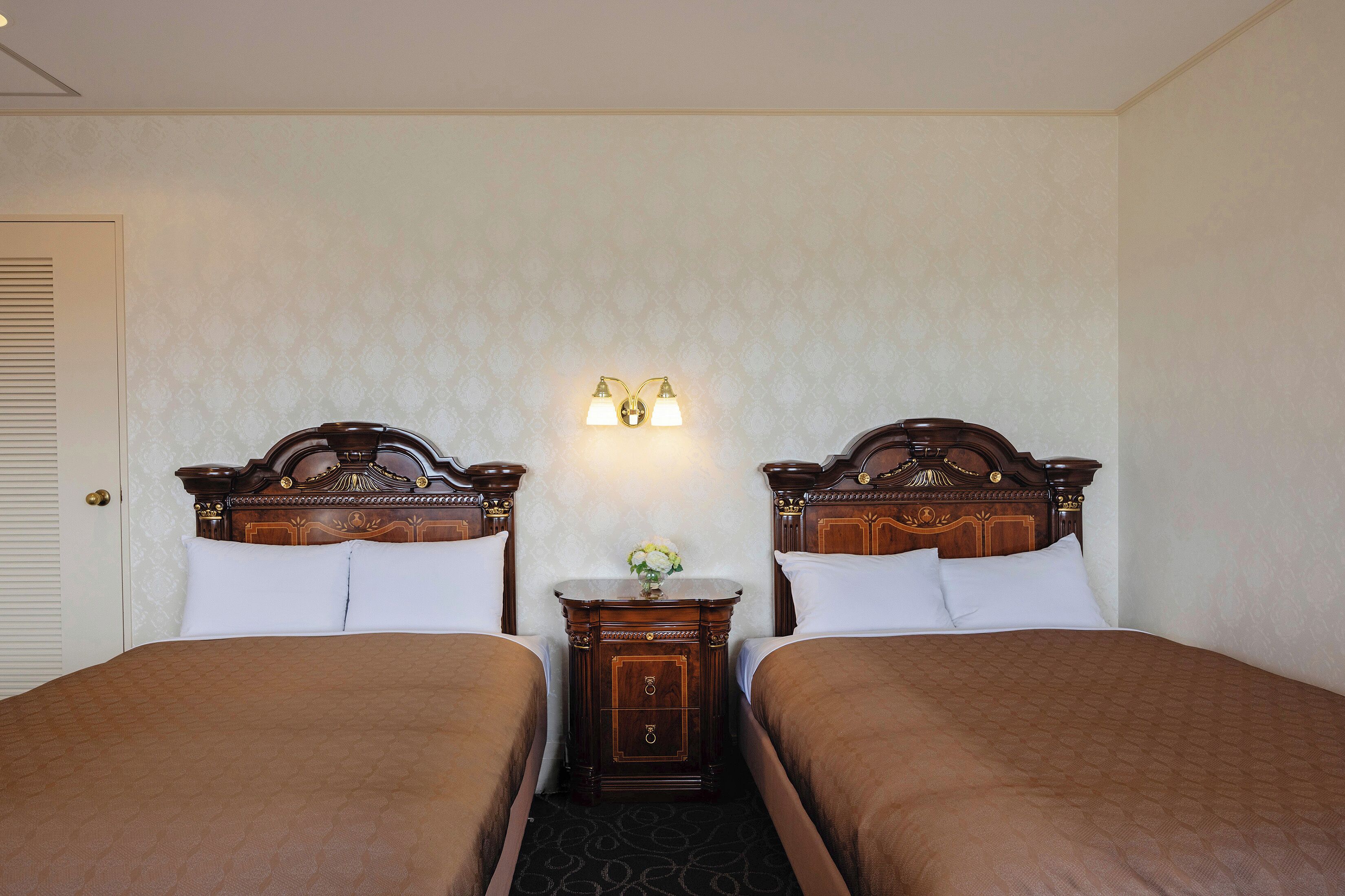 A hotel room with two single beds, a nightstand with a vase of flowers, and a wall-mounted light fixture.