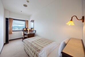 A hotel room with a bed, a desk, a chair, a window with curtains, a mirror, and a wall-mounted lamp.