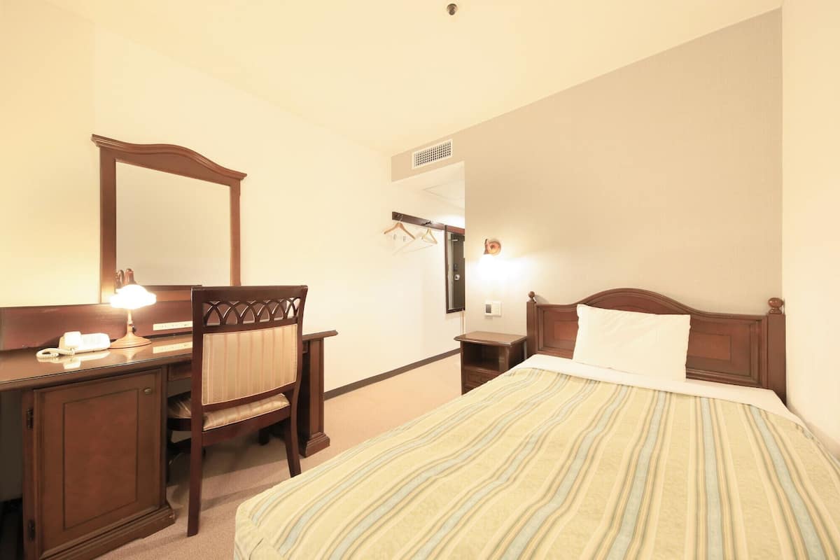 Una habitación de hotel con cama, escritorio con teléfono, silla, espejo y calentador de pared.