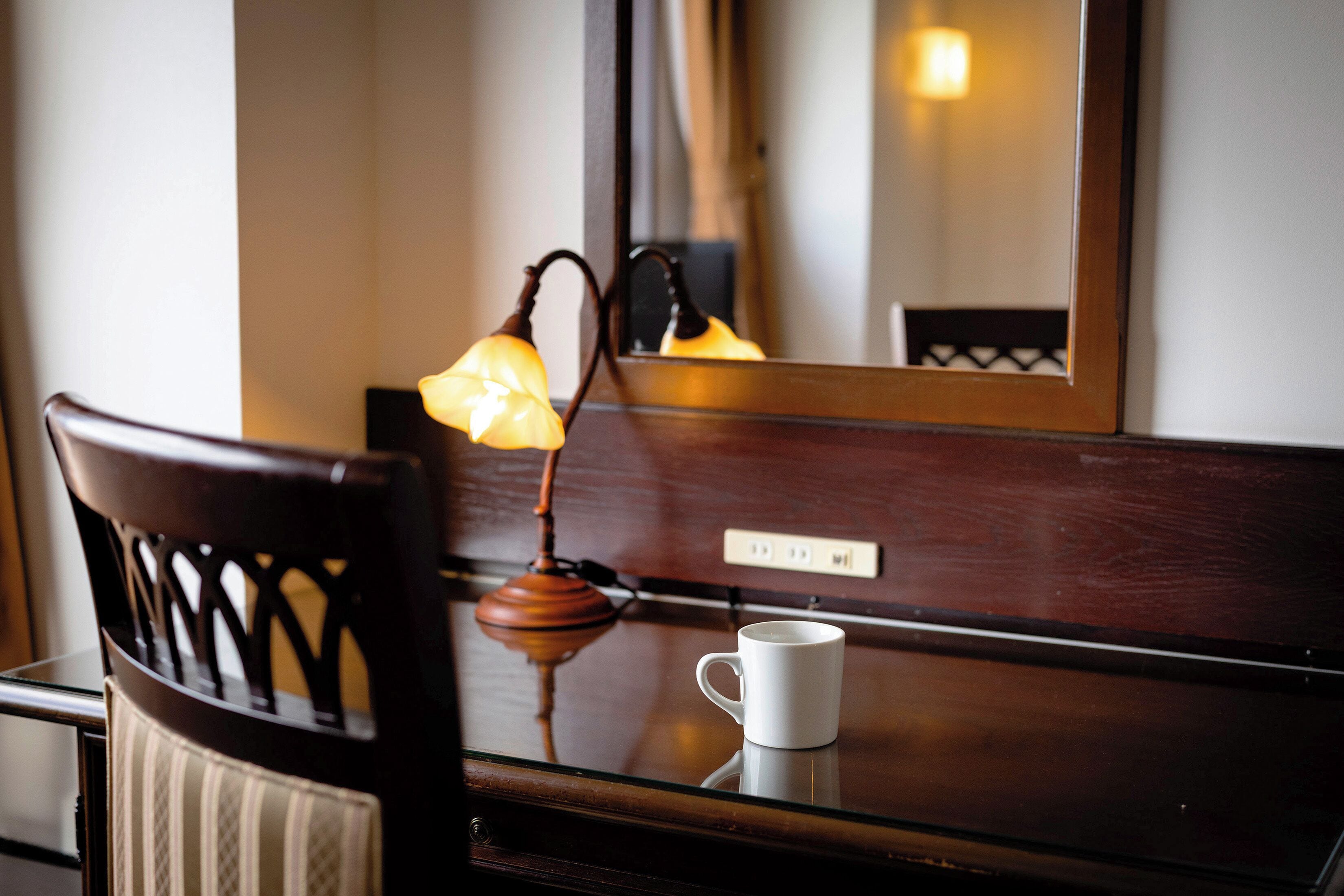 A wooden chair with a cushion, a white mug on a reflective surface, and a wall-mounted lamp with a visible switch.