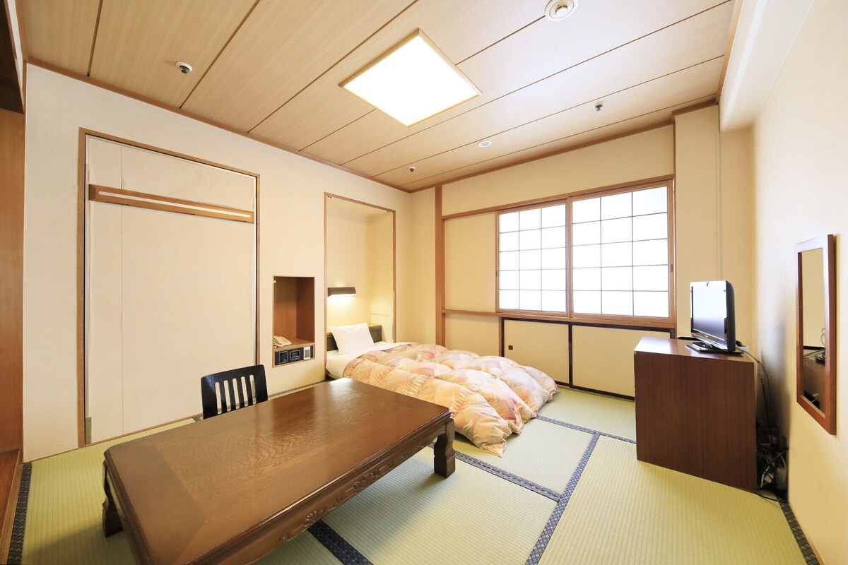 Una habitación tradicional japonesa con suelo de tatami, una cama con edredón rosa y blanco, un escritorio de madera con televisión y una puerta corredera.