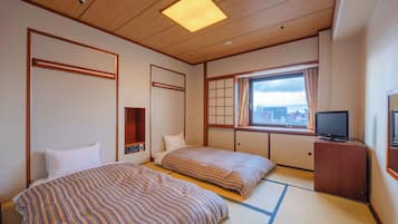 A traditional Japanese-style hotel room with tatami flooring, two beds with striped bedding, a television on a wooden stand, and a window with a view of the city.