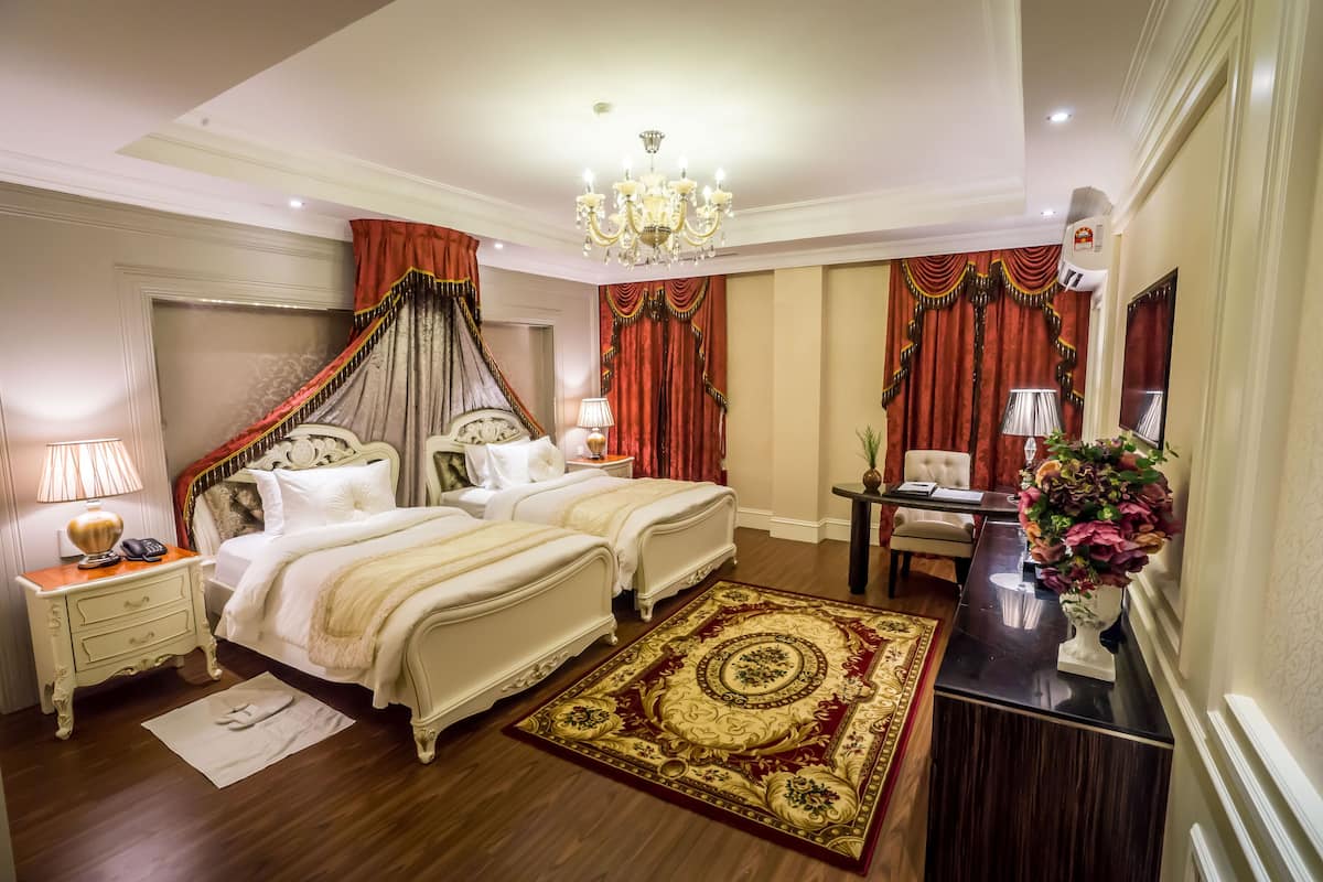 Ein luxuriöses Schlafzimmer mit zwei Betten, einer Kristallleuchter, einem Schreibtisch mit Blumenvase und einem Teppich mit roten und goldenen Mustern.