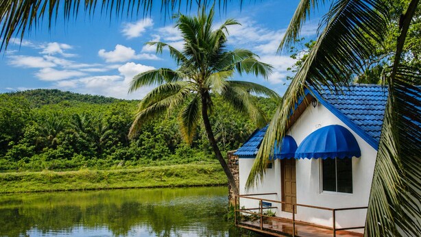 A house with a blue roof and white walls, surrounded by a lake and palm trees. - The Orchard Resort & Spa Melaka (Alor Gajah)