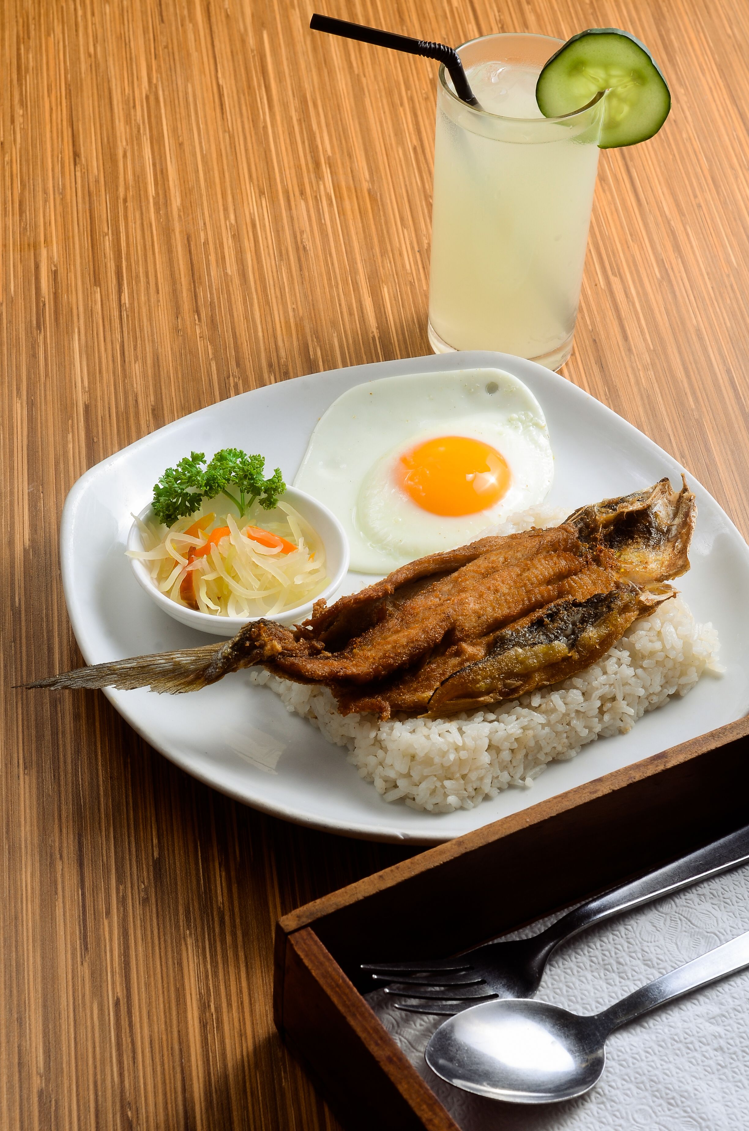 A plate with rice, fried fish, a fried egg, and a cucumber slice on a wooden table.