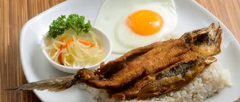 A plate with rice, fried fish, a fried egg, and a cucumber slice on a wooden table.