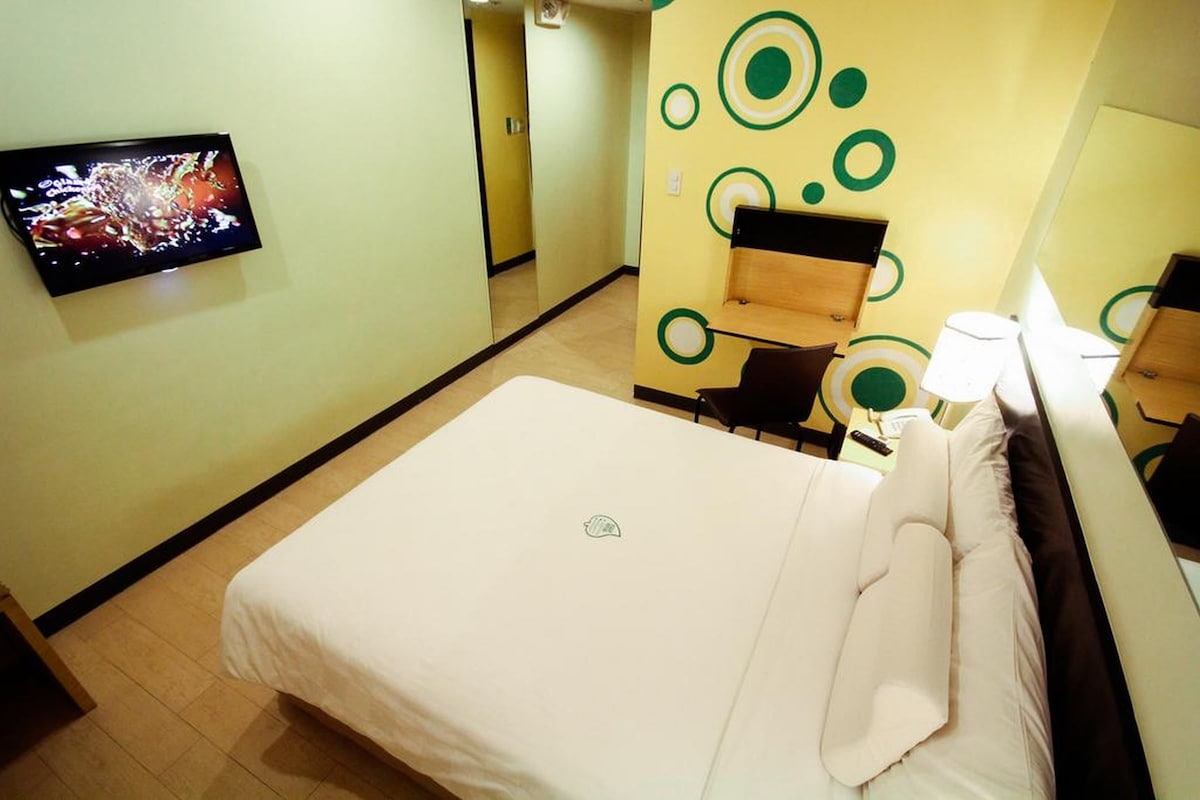 A hotel room with a bed, a chair, a desk with a lamp, and a television on the wall.