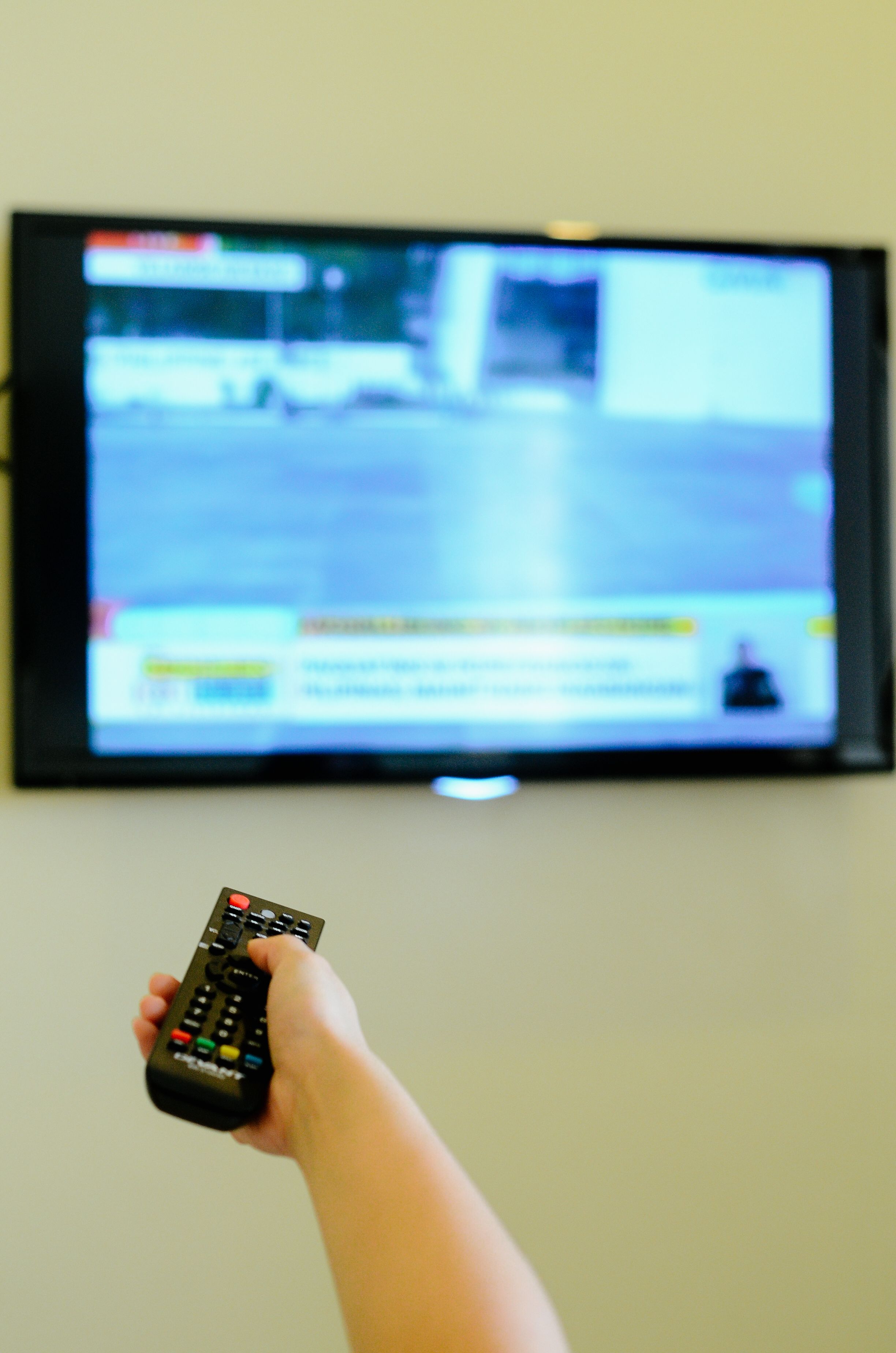 A hand holding a remote control in front of a television displaying a news channel.