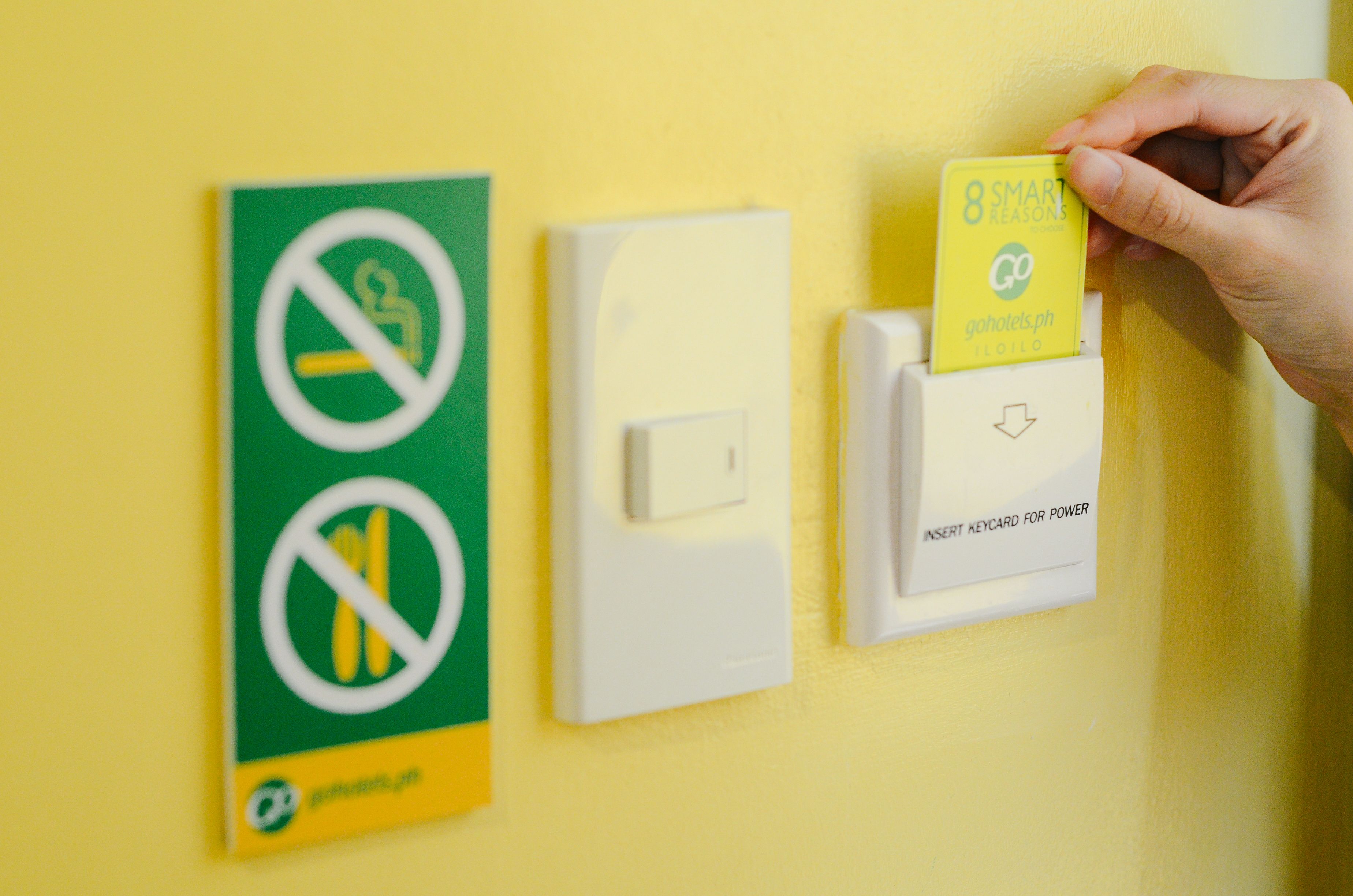 A hand inserting a card into a slot on a yellow wall, next to a light switch and a sign with a prohibition symbol.