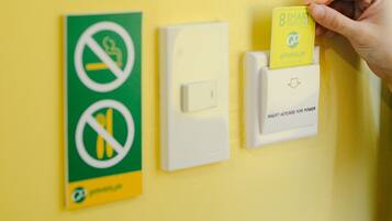 Una mano insertando una tarjeta en una ranura en una pared amarilla, cerca de un interruptor de luz y un cartel con un símbolo de prohibición.