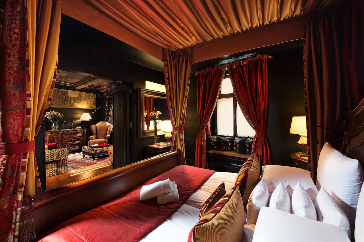 Um quarto espaçoso com uma cama grande, um espaço de estar com uma poltrona e uma mesa, e vista para a cidade pela janela.