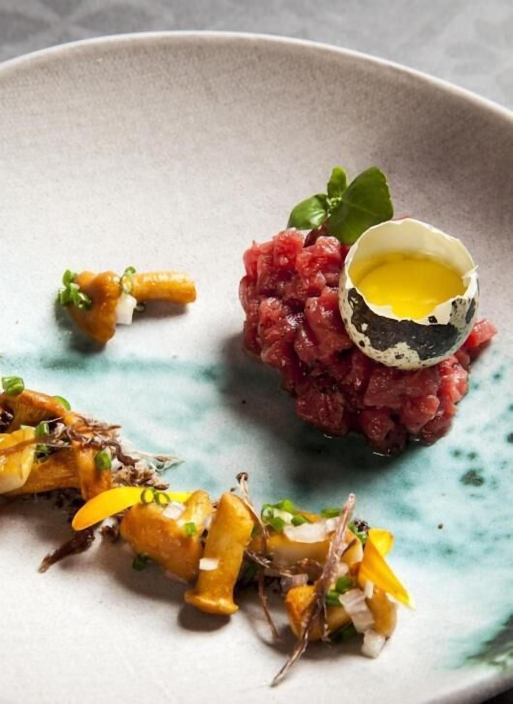 A plate with a dish of raw meat, a cracked egg, and some garnishes.