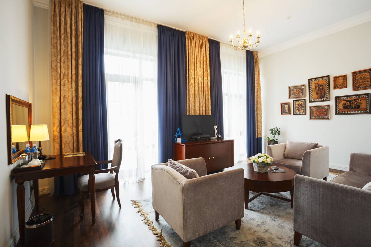 Habitación de hotel con un escritorio de madera, dos sillones, un sofá, un televisor sobre un mueble de madera y una araña.