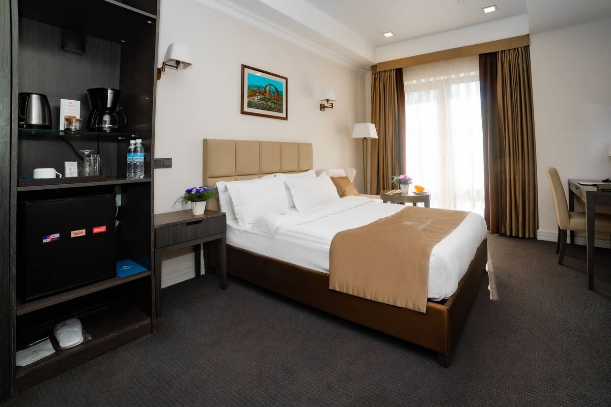 Habitación de hotel con cama, escritorio, silla, cafetera, botella de agua y un cuadro en la pared.