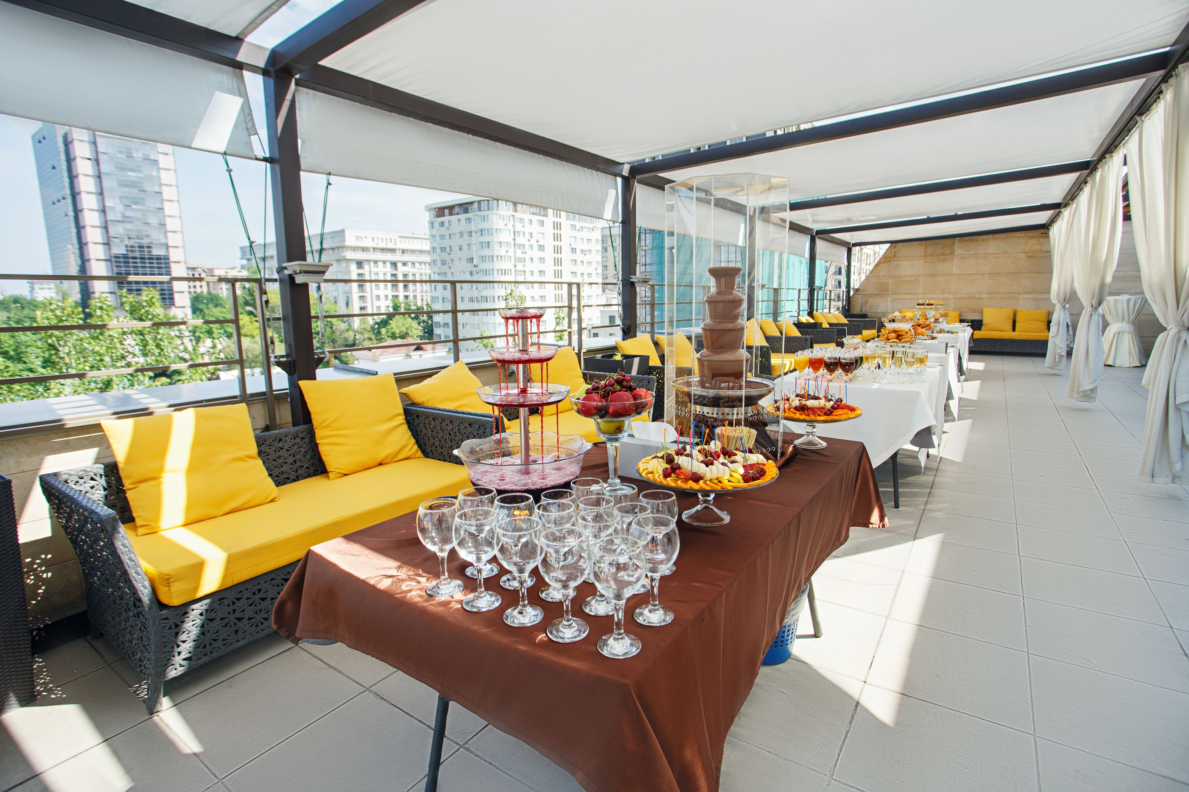 Un área de comedor al aire libre con una mesa larga dispuesta para una comida, rodeada de asientos acolchados amarillos y vistas a la ciudad.