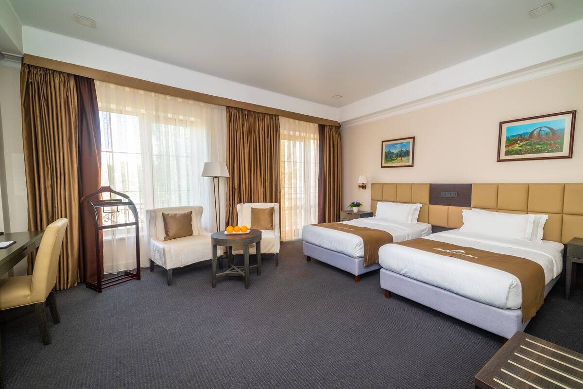 Habitación de hotel con dos camas, un escritorio, una silla, una mesita con fruta y dos cuadros en la pared.