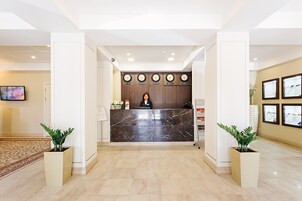 Zona de recepción de un hotel con un mostrador de mármol, una recepcionista y una pared con relojes que muestran la hora en diferentes ciudades.