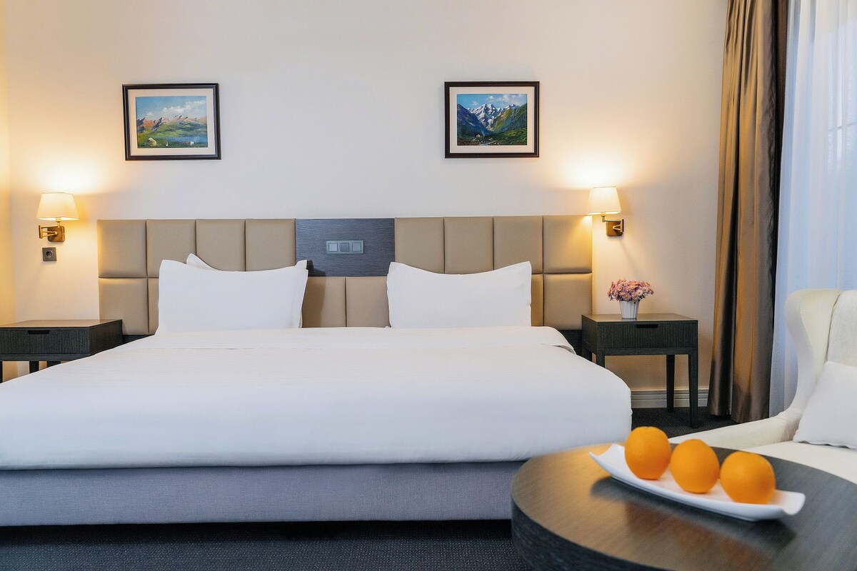 Une chambre d’hôtel avec un grand lit, deux lampes fixées au mur, des tableaux encadrés, une table de chevet avec un vase et une chaise.
