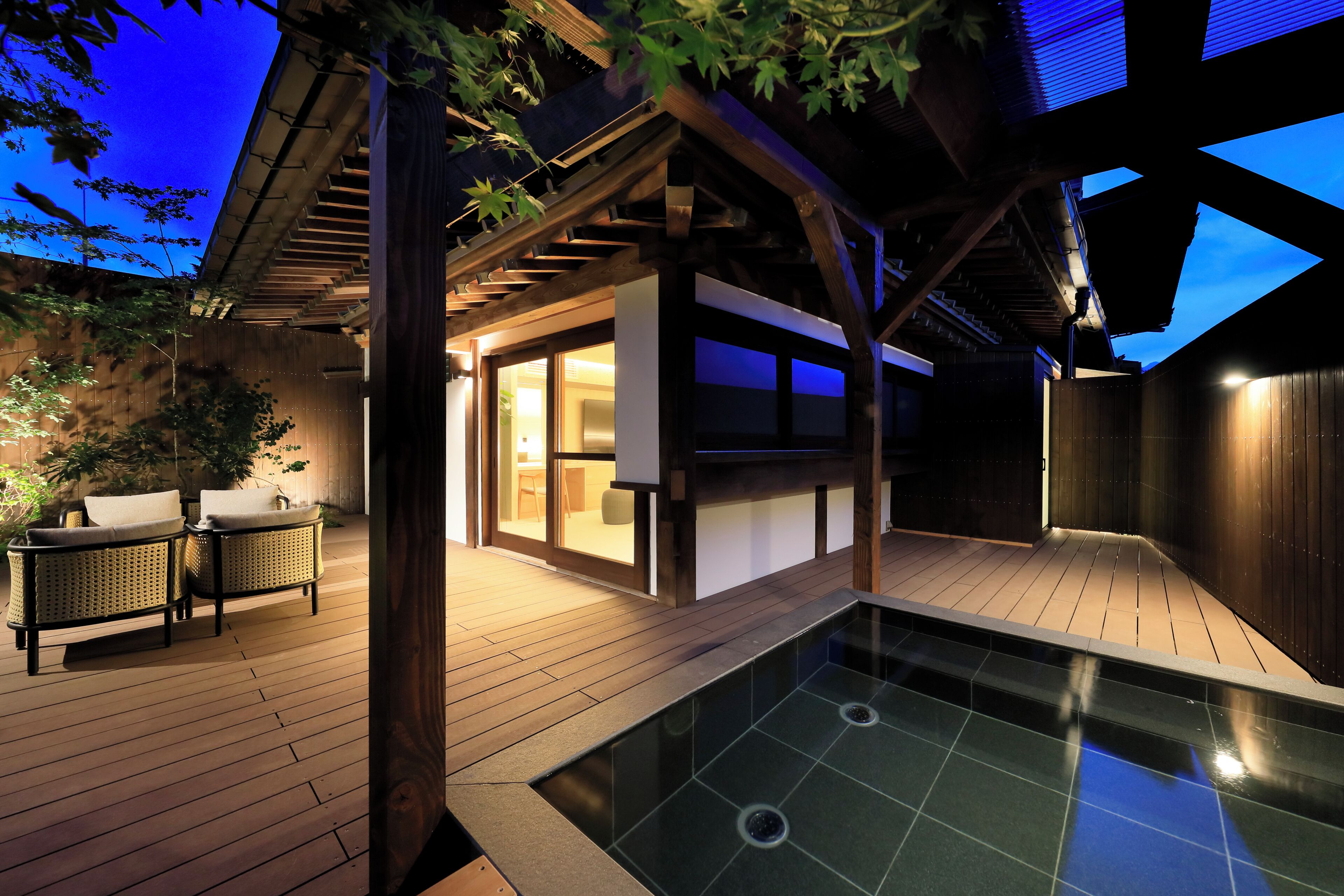 A modern outdoor pool area with a wooden deck, a seating area with cushions, and a glass-enclosed structure.