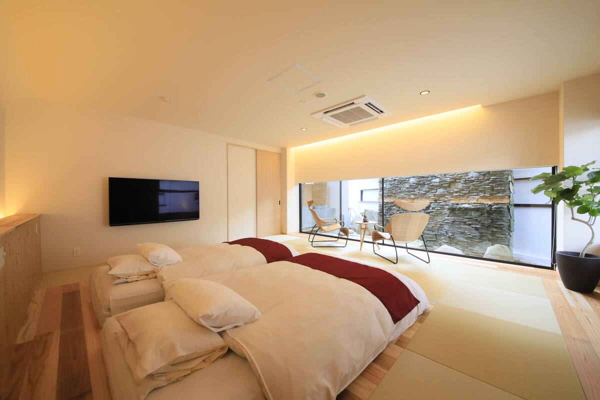 Ein modernes Schlafzimmer mit einem großen Bett, einem Fernseher und Blick auf eine Steinwand sowie einen Sitzbereich im Freien.