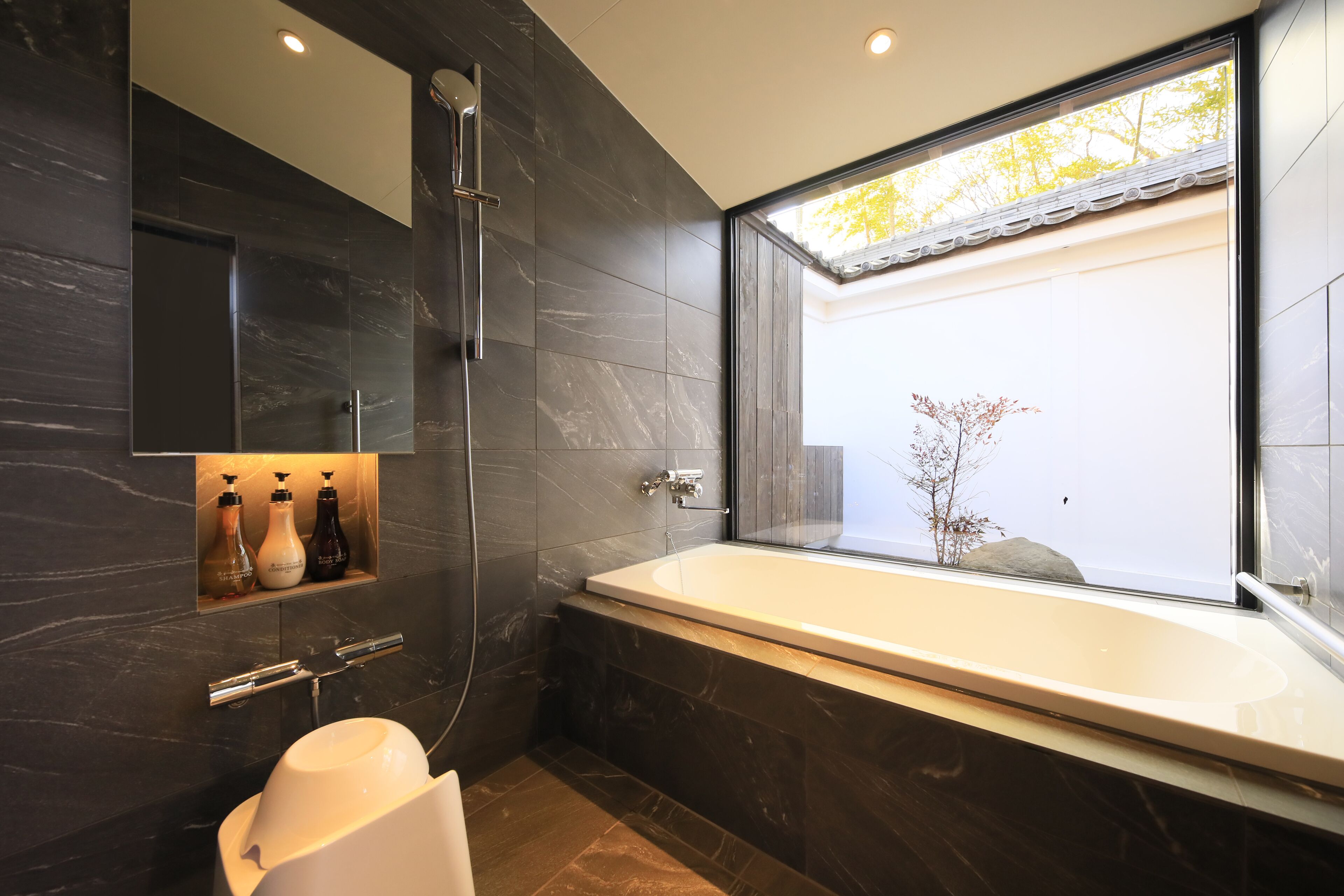 A modern bathroom with a large bathtub, dark marble walls, and a window with a view of trees.