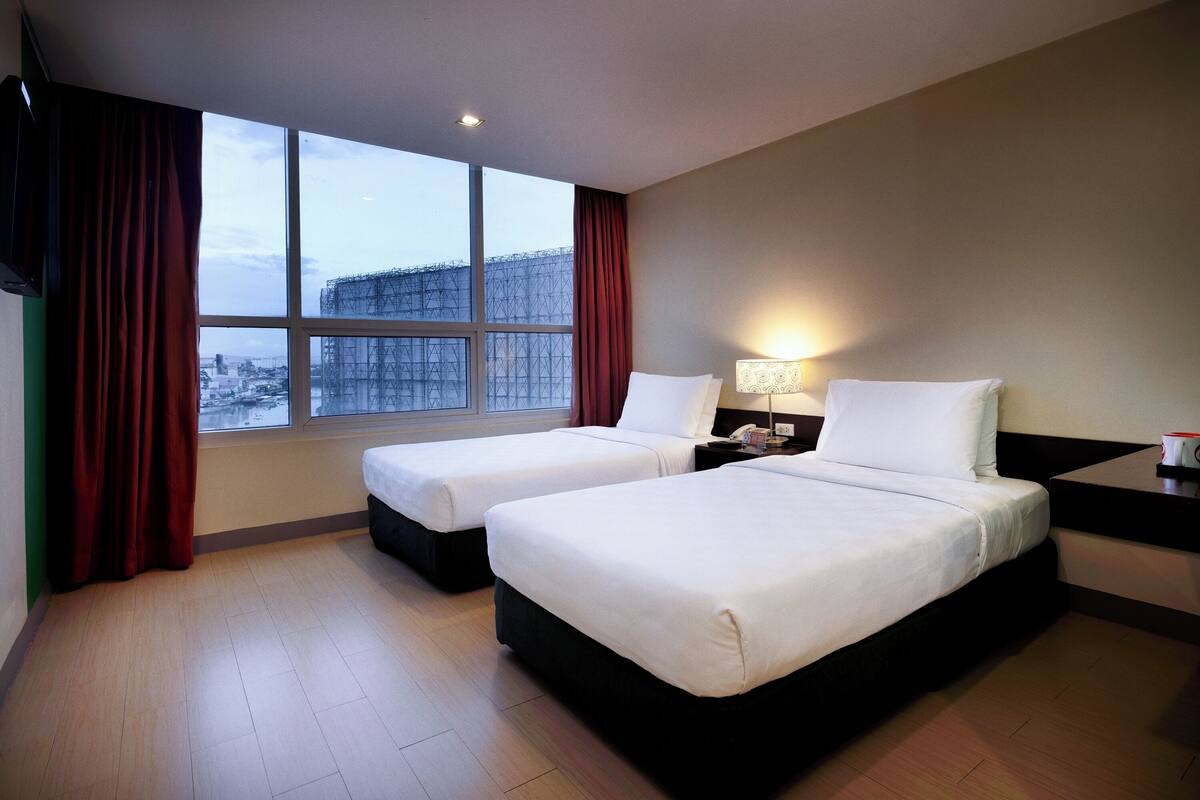 A hotel room with two beds, a nightstand with a lamp, a window with red curtains, and a view of a cityscape.