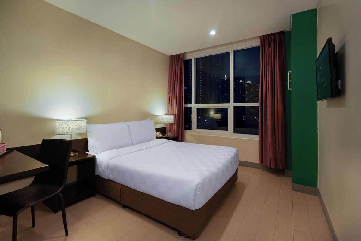 A hotel room with a bed, a desk with a chair, a TV mounted on the wall, and a window with curtains.