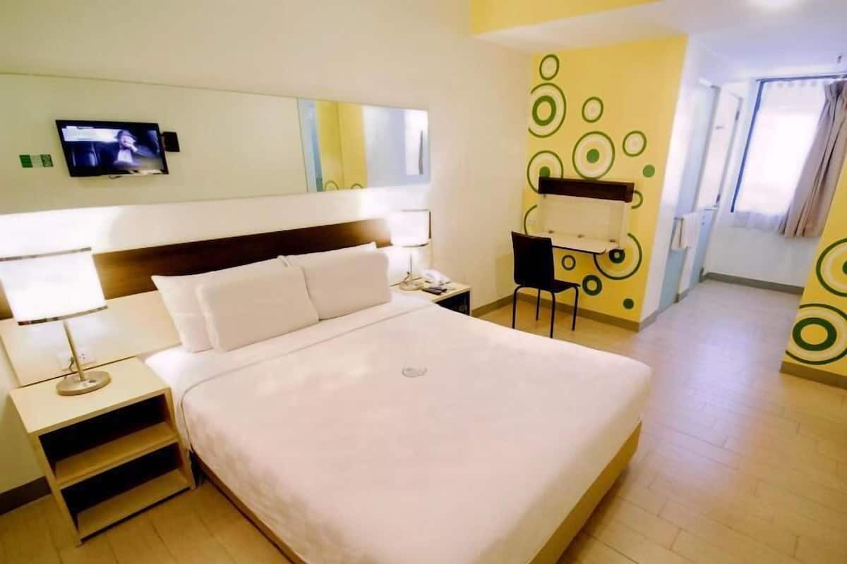 A hotel room with a bed, bedside table, lamp, TV, desk, chair, and a mirror.