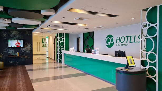 A hotel reception area with a green reception desk, a digital screen displaying a person, and a sign that reads "GO HOTELS OTIS - MANILA". - Go Hotels Otis - Manila (Manila)