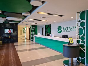 Ein Hotel-Rezeptionsbereich mit einem grünen Empfangstresen, einem digitalen Bildschirm, der eine Person zeigt, und einem Schild mit der Aufschrift 'GO HOTELS OTIS - MANILA'.
