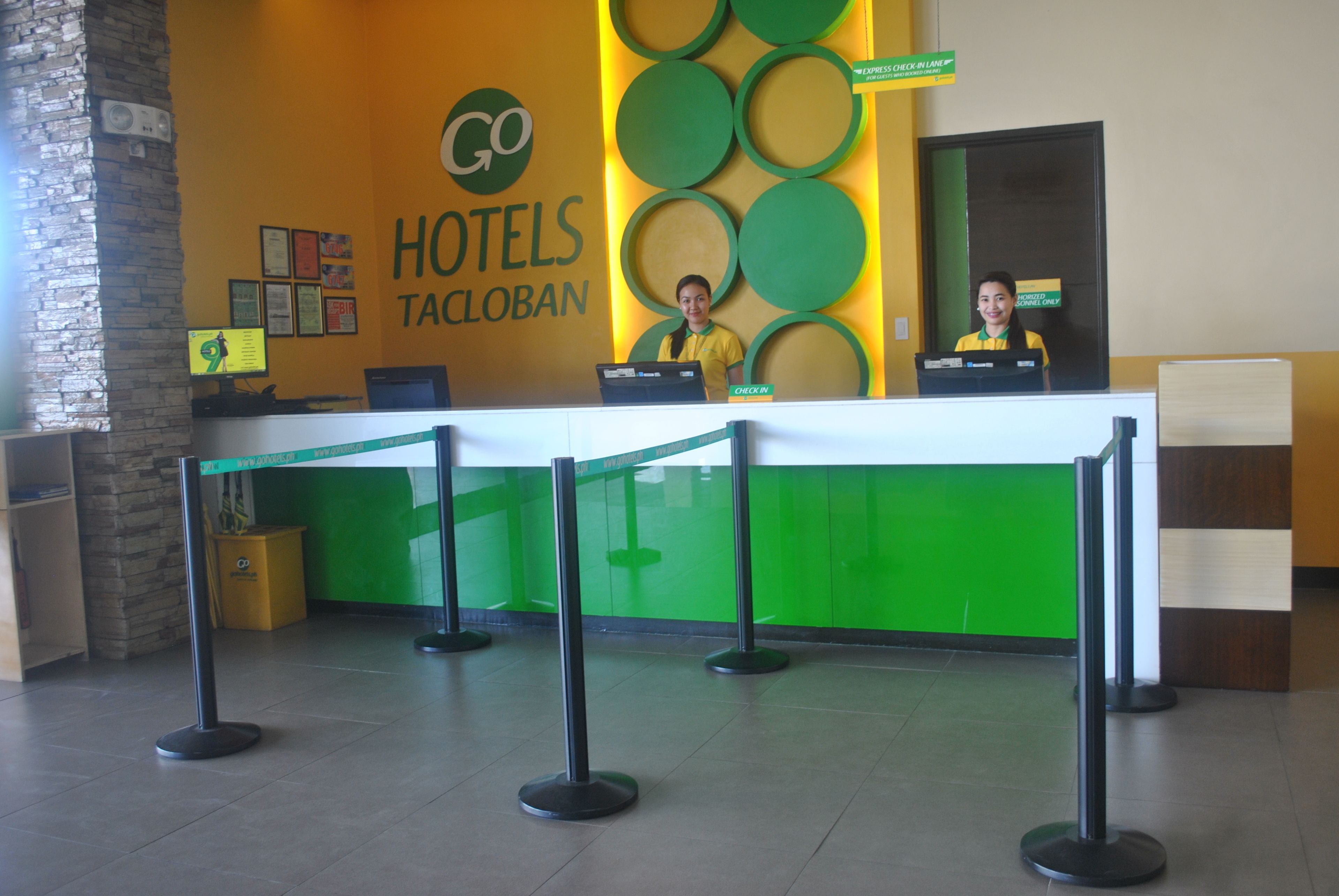 Un mostrador de recepción de hotel con una decoración en tonos verde y amarillo, dos recepcionistas y un mostrador de registro.