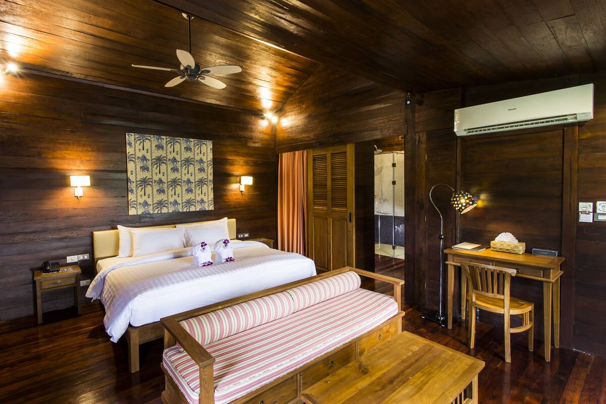 A wooden-floored room with a bed, bedside table, bench, desk, chair, and an air conditioning unit.