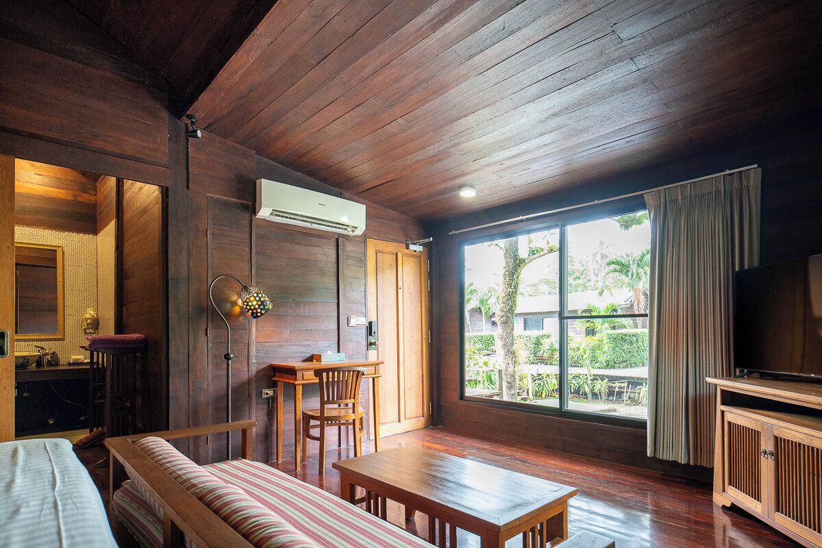 Habitación con suelo de madera, una cama, un sofá, una mesa, un televisor y un ventanal con vistas a la vegetación.