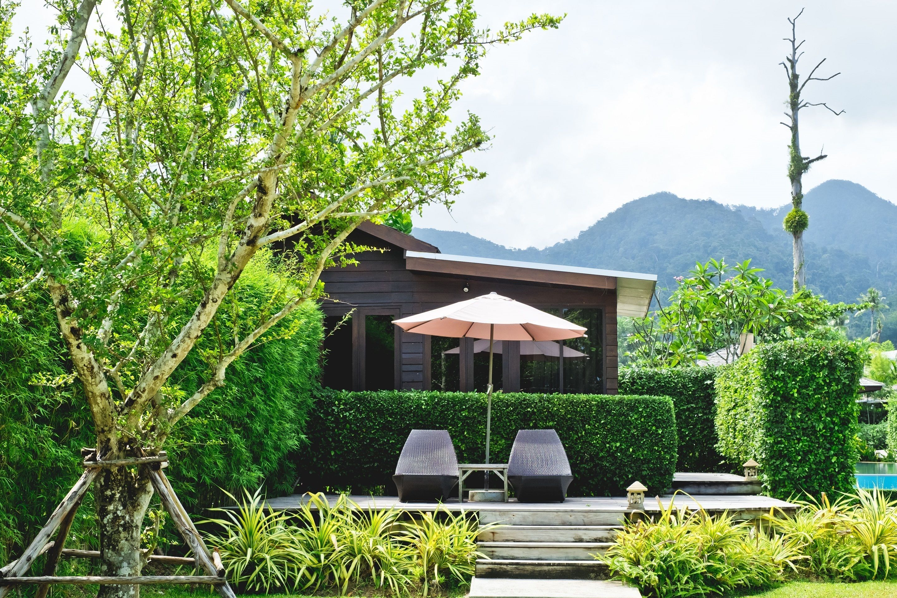 A modern outdoor seating area with a table and chairs under a large umbrella, surrounded by lush greenery and a mountain backdrop.