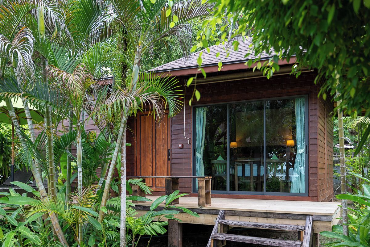 A wooden house with large glass windows, surrounded by lush tropical vegetation.